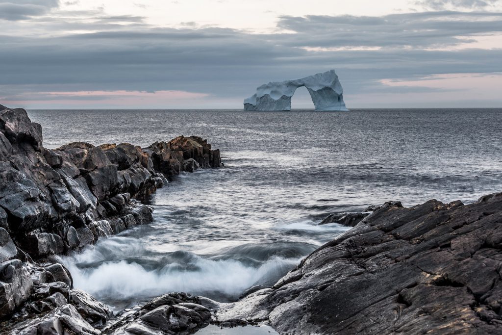 An arched iceberg is seen a short distance from a rocky shore with waves