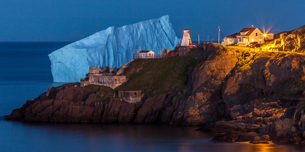 A massive iceberg at blue hour behind a peninsula with a lighthouse