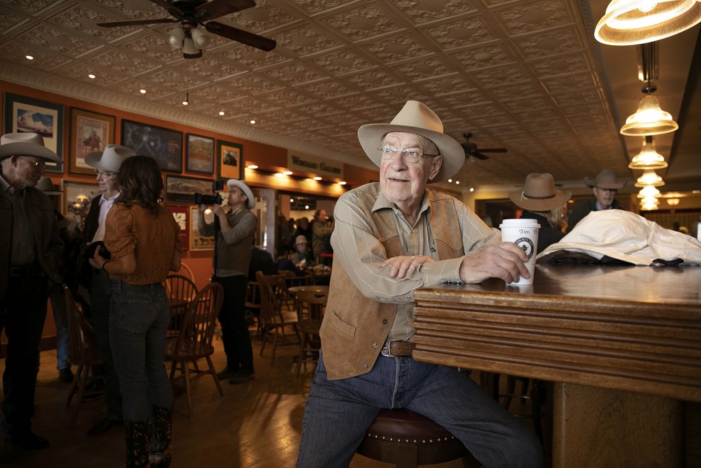 An elderly man in a cowboy hat drinks coffee while sitting at a bar