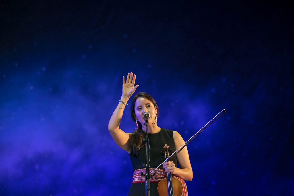 A Métis woman wearing a black dress with a ceinteur fléchée and holding a fiddle waves to the audience