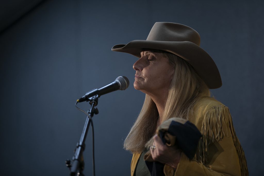 A blonde woman wearing a cowboy hat closes her eyes as she speaks into a microphone