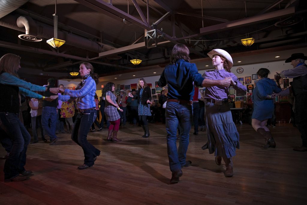 Couples in traditional western dress dance in a large room with wooden floors and rafters