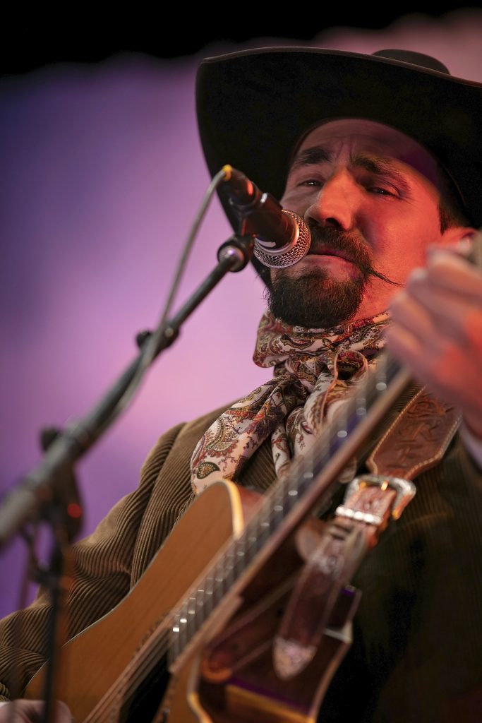 A man in a cowboy hat with goatee and handlebar moustache performs with a guitar