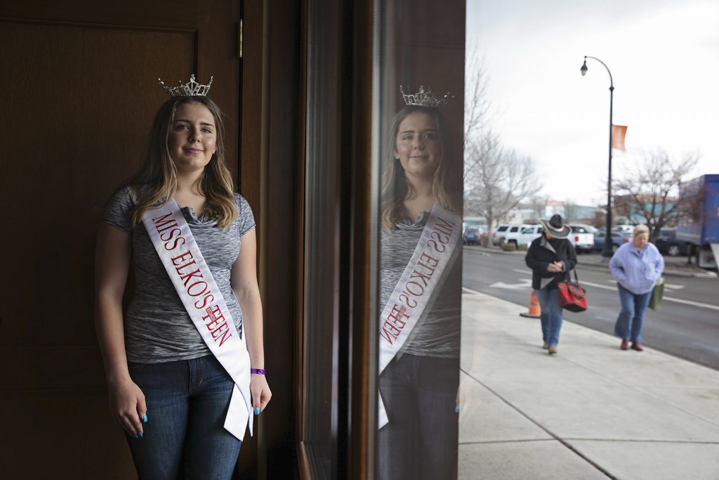 A teenage girl poses in front of a window wearing a tiara and a sash that reads
