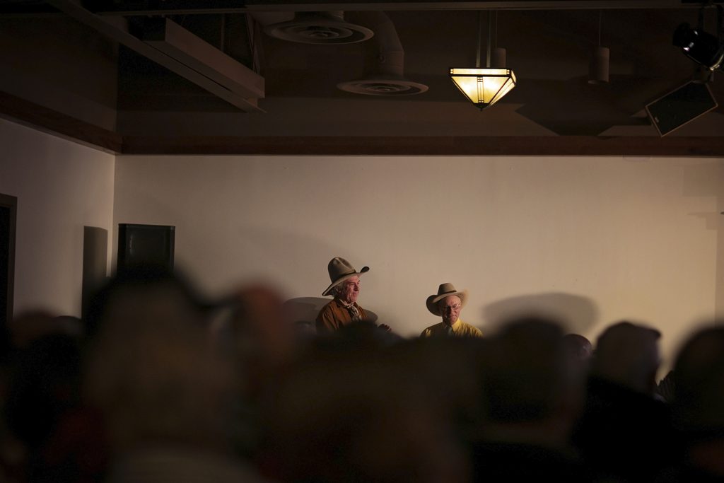 Two elderly gentlemen in cowboy hats are visible behind the blurred heads of an attentive audience