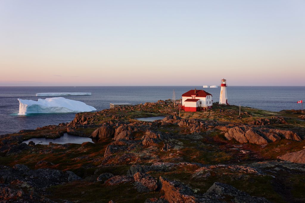 A large iceberg is seen off the rocky coast of an island with a lighthouse