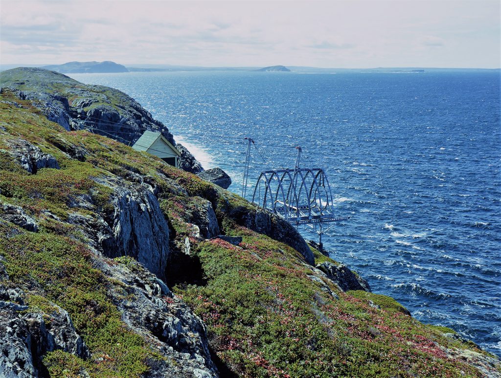 A metal frame anchored in a cliff juts out over a blue ocean. This is a whale-watching pod under construction.