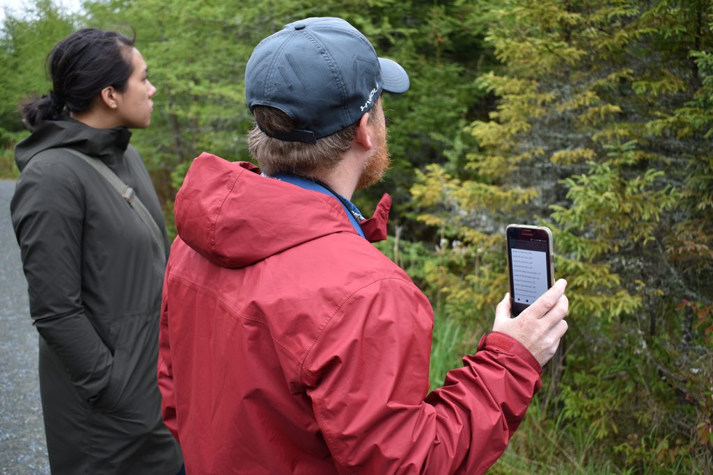 A man in a red rain jacket holds up a phone in front of some trees, playing a bird call