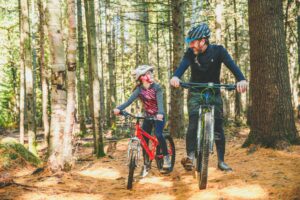 Un père et sa fille, souriants, font du vélo sur un sentier dans la forêt.