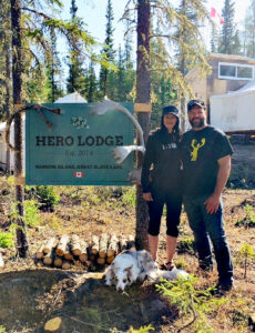Couple dans une forêt devant un chalet où à leurs pieds, repose un chien.