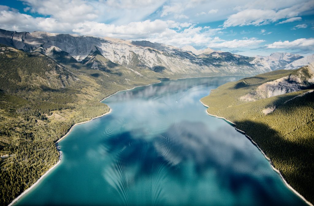 Aerial view of turquoise lake surrounded by forests and expansive mountain range.